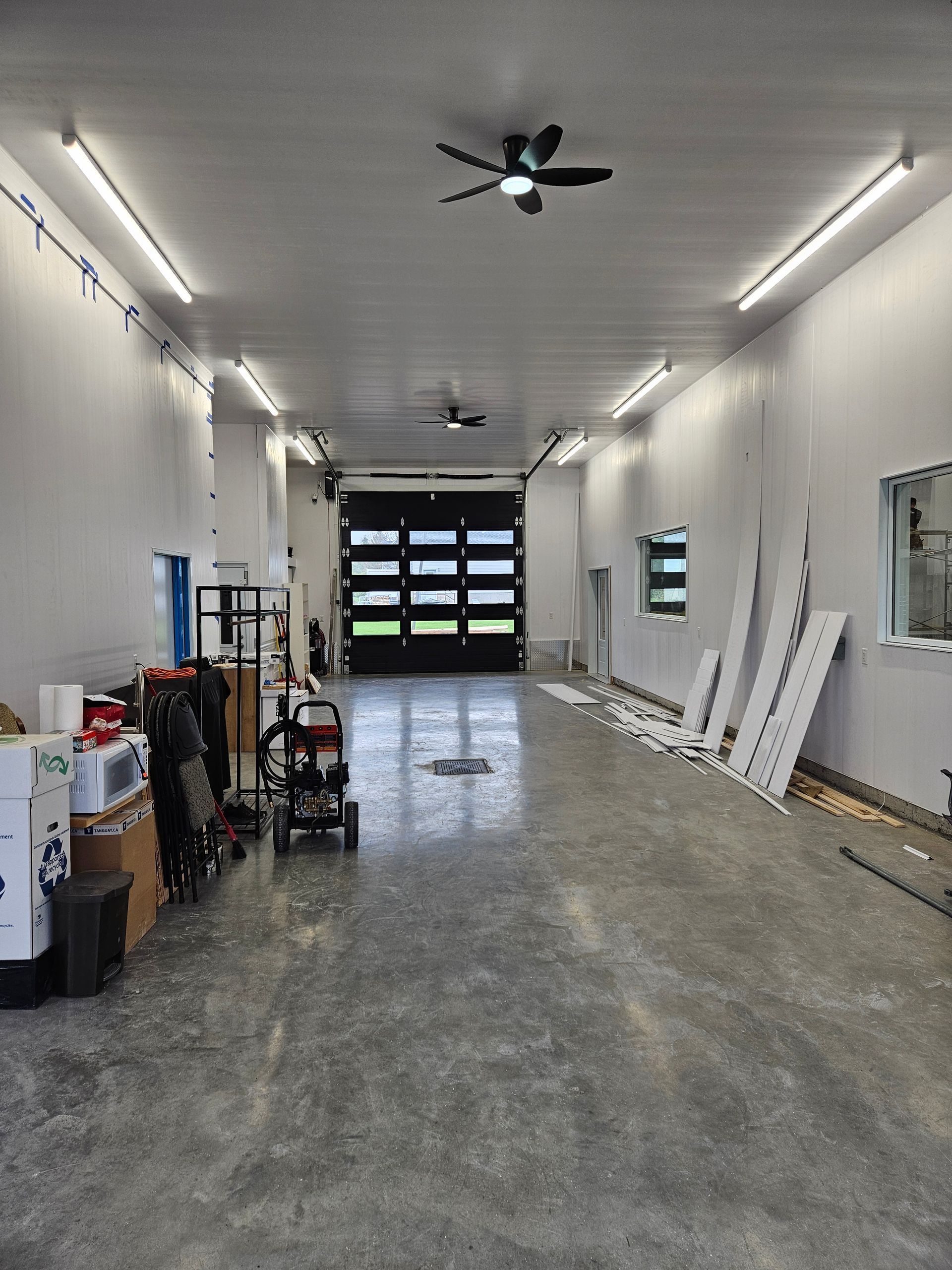 Un garage vide et spacieux avec un sol en béton poli, des murs blancs, un éclairage au plafond et une grande porte de garage noire.