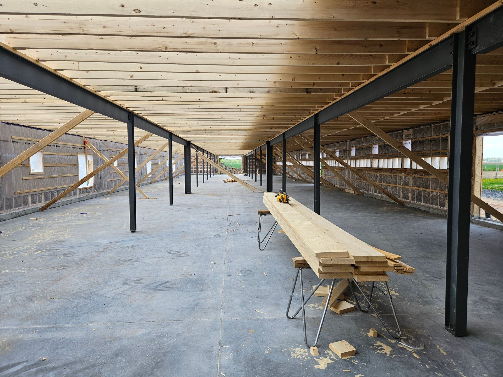 Intérieur d'un bâtiment en construction présentant des solives de plafond en bois apparentes, des poutres de soutien en acier et une table sur tréteaux.