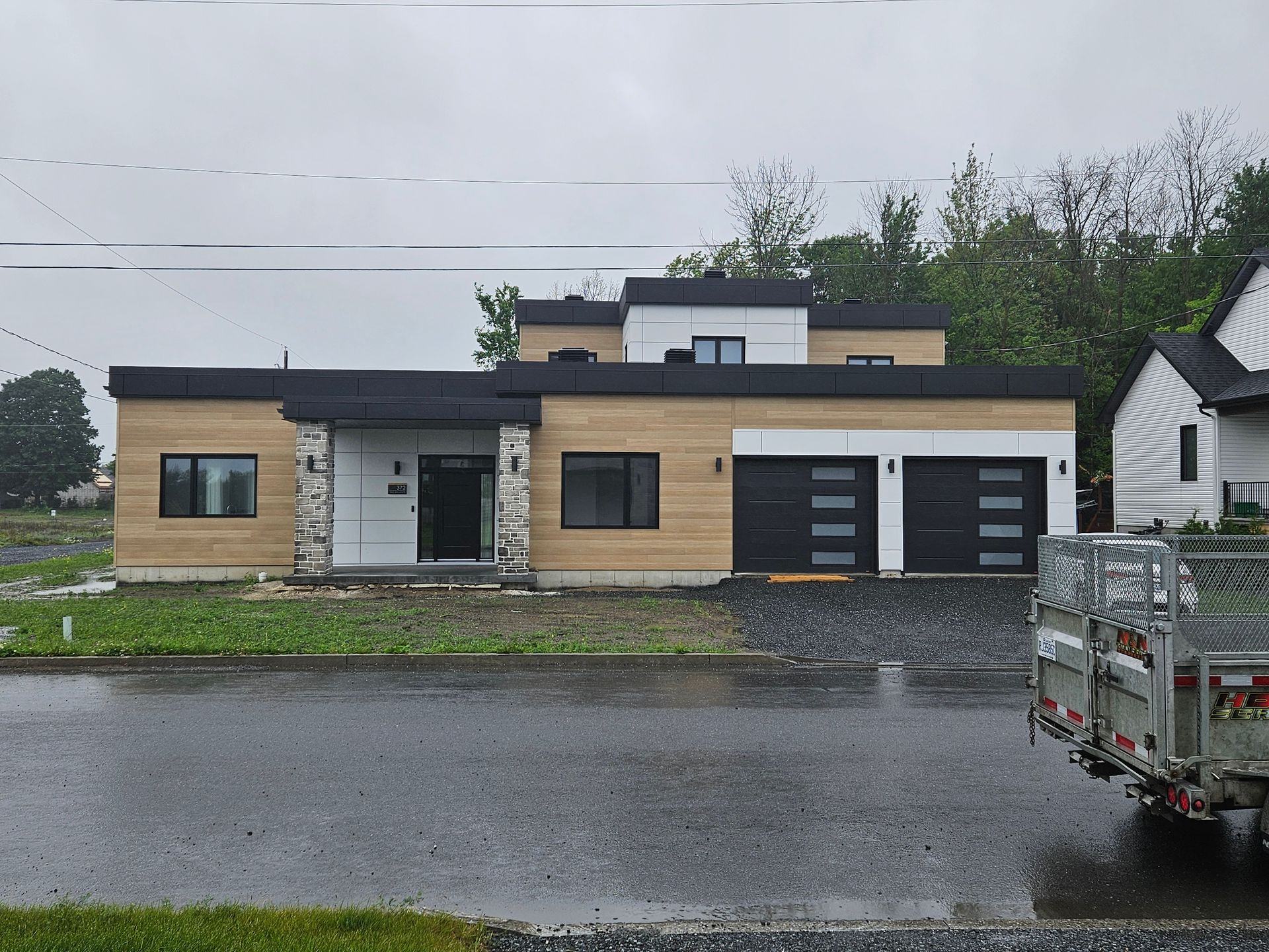 Une maison moderne à toit plat avec un revêtement extérieur imitation bois, des éléments en pierre, deux portes de garage foncées et un étage partiel.