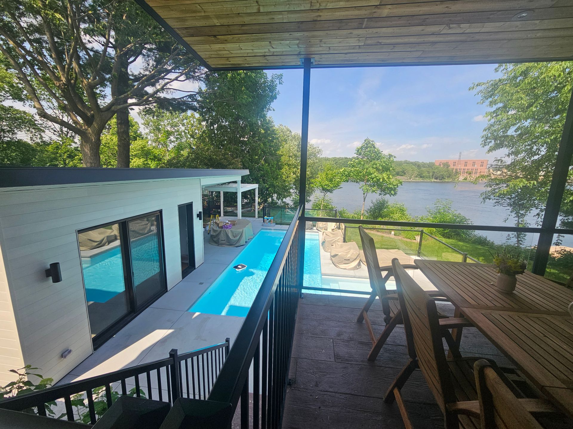 Une terrasse extérieure en bois avec mobilier de jardin donne sur une maison blanche moderne avec piscine et rivière.
