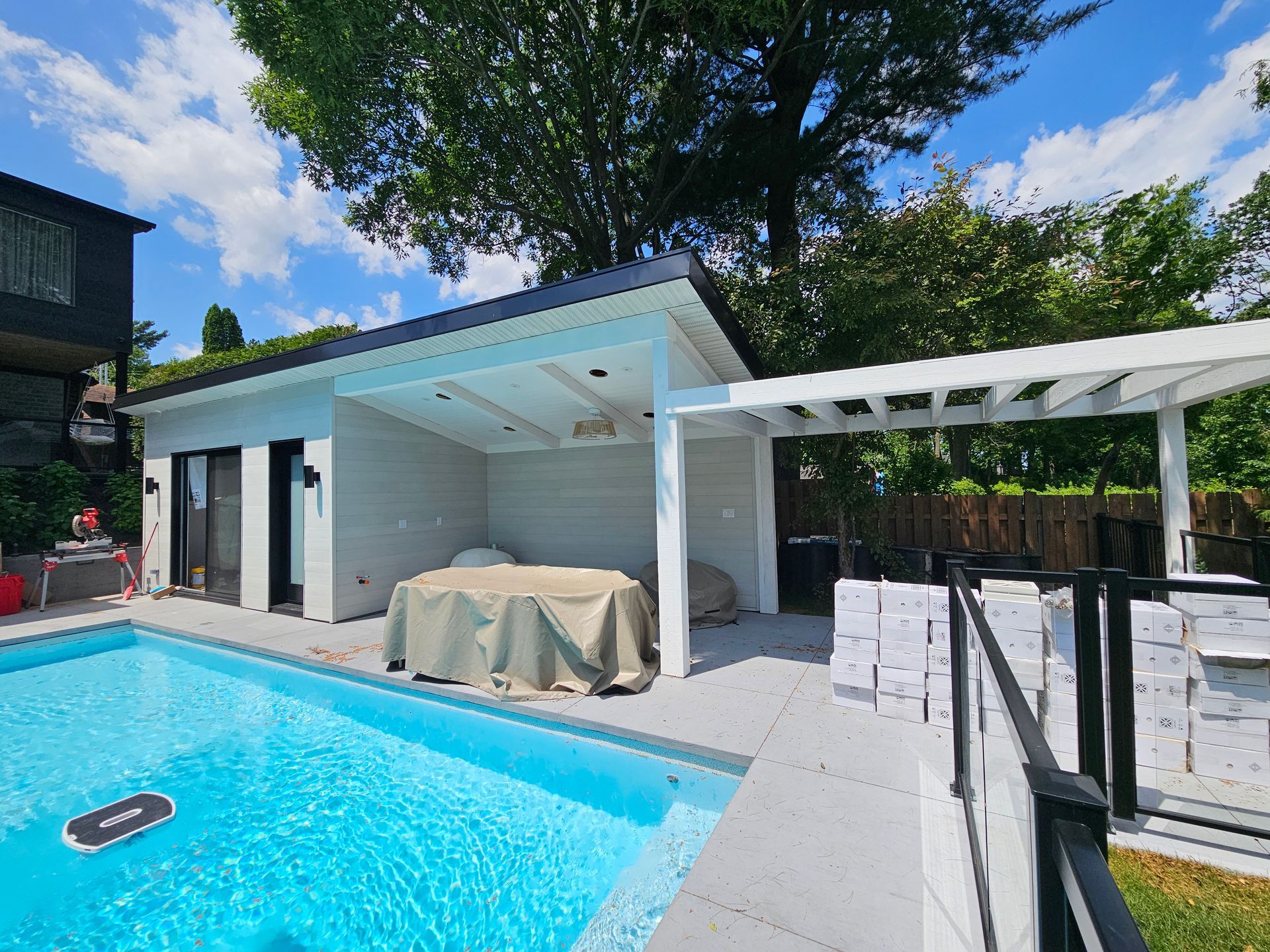 Un pool house blanc avec terrasse couverte et pergola, situé à côté d'une piscine bleu vif.