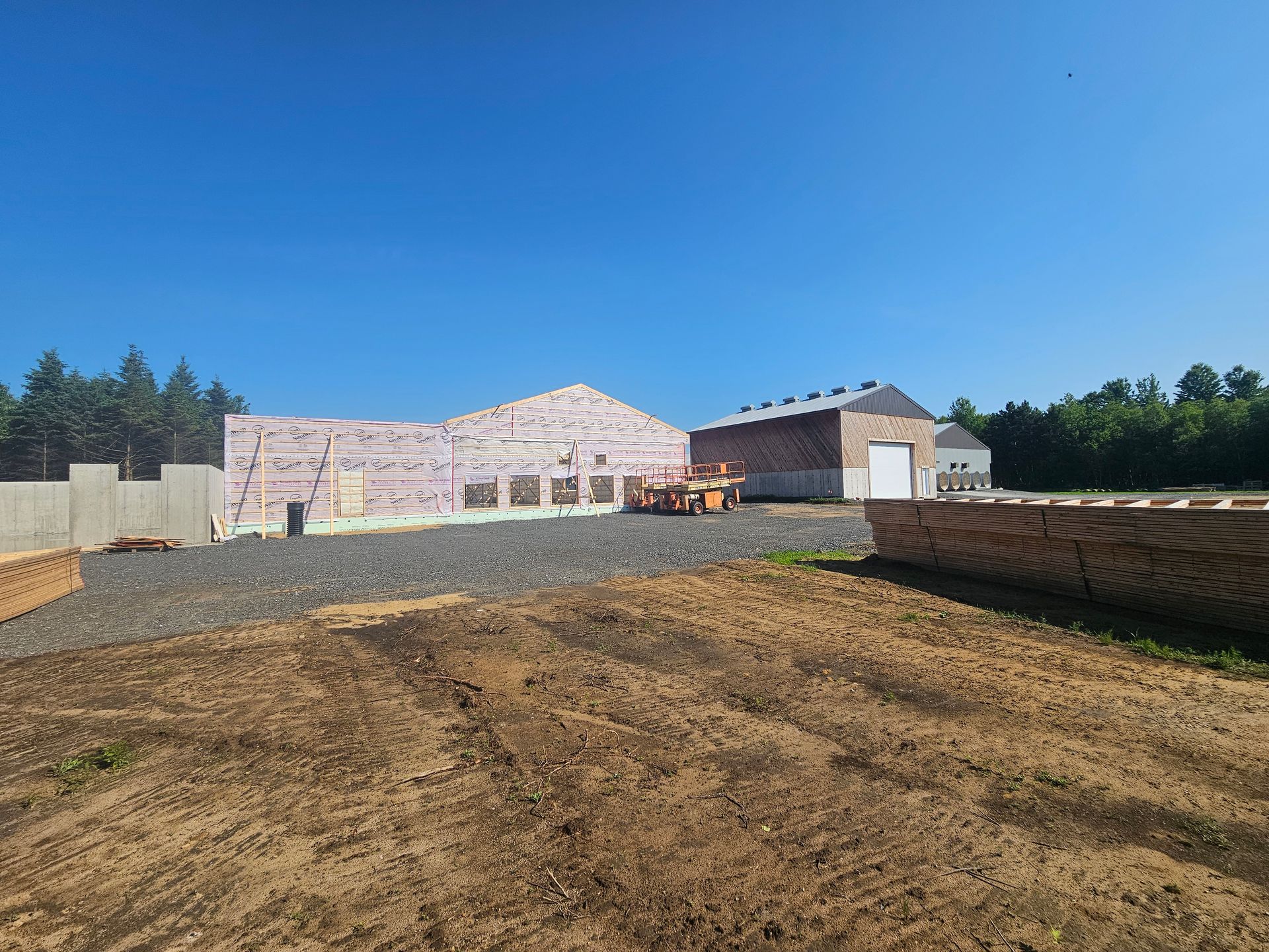 Un chantier avec une structure partiellement construite, un sol gravillonné, des piles de bois et un ciel bleu dégagé.