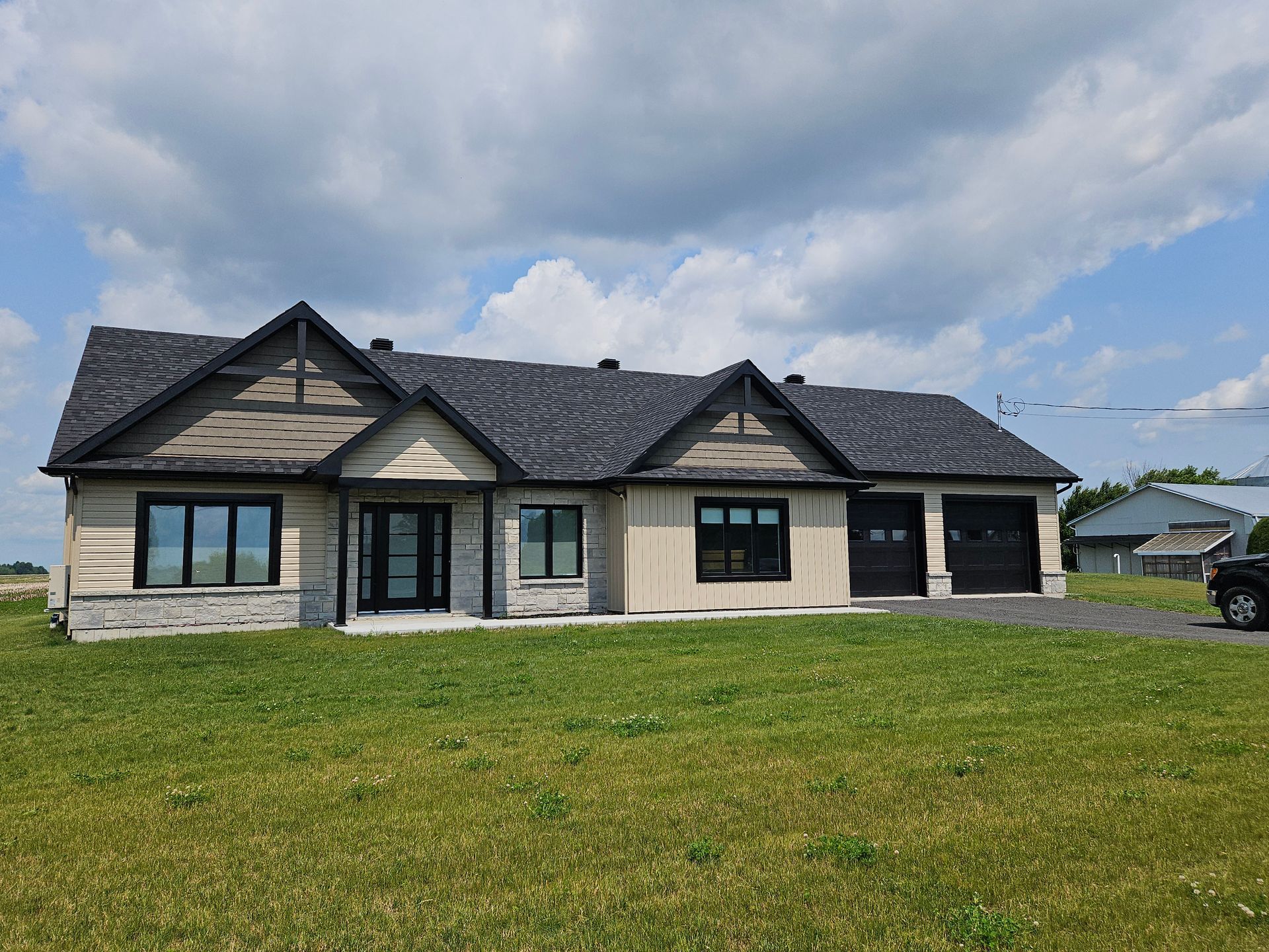 Une maison moderne de plain-pied, avec un revêtement extérieur beige, des éléments en pierre, des boiseries foncées et un garage double, se dresse sur un terrain gazonné.