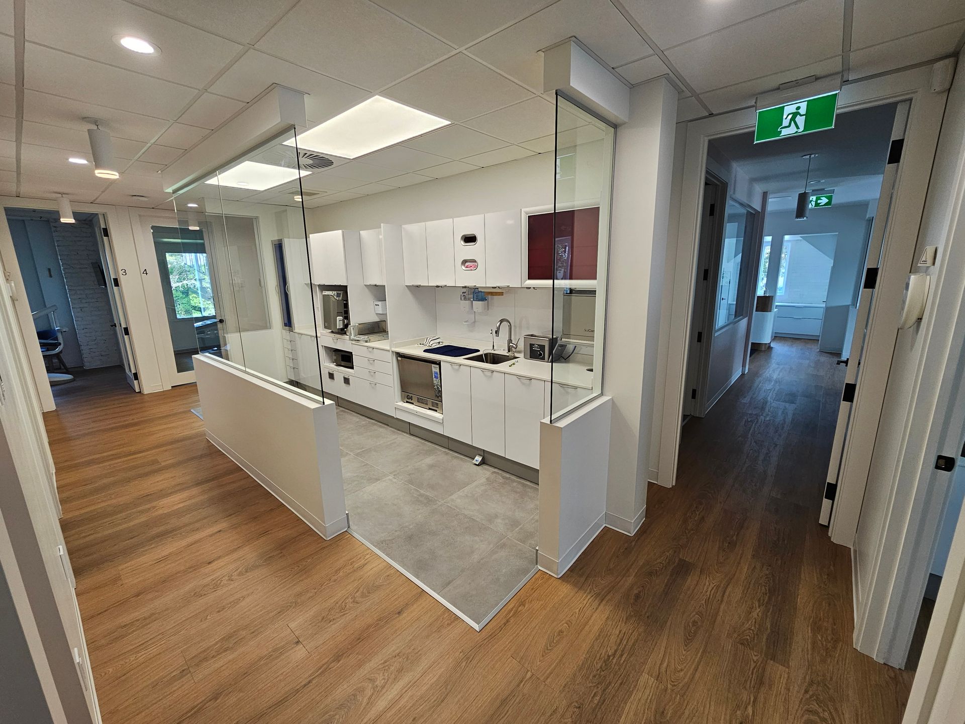Une cuisine de bureau moderne avec des armoires blanches, un parquet clair et une cloison vitrée, vue depuis un couloir adjacent.