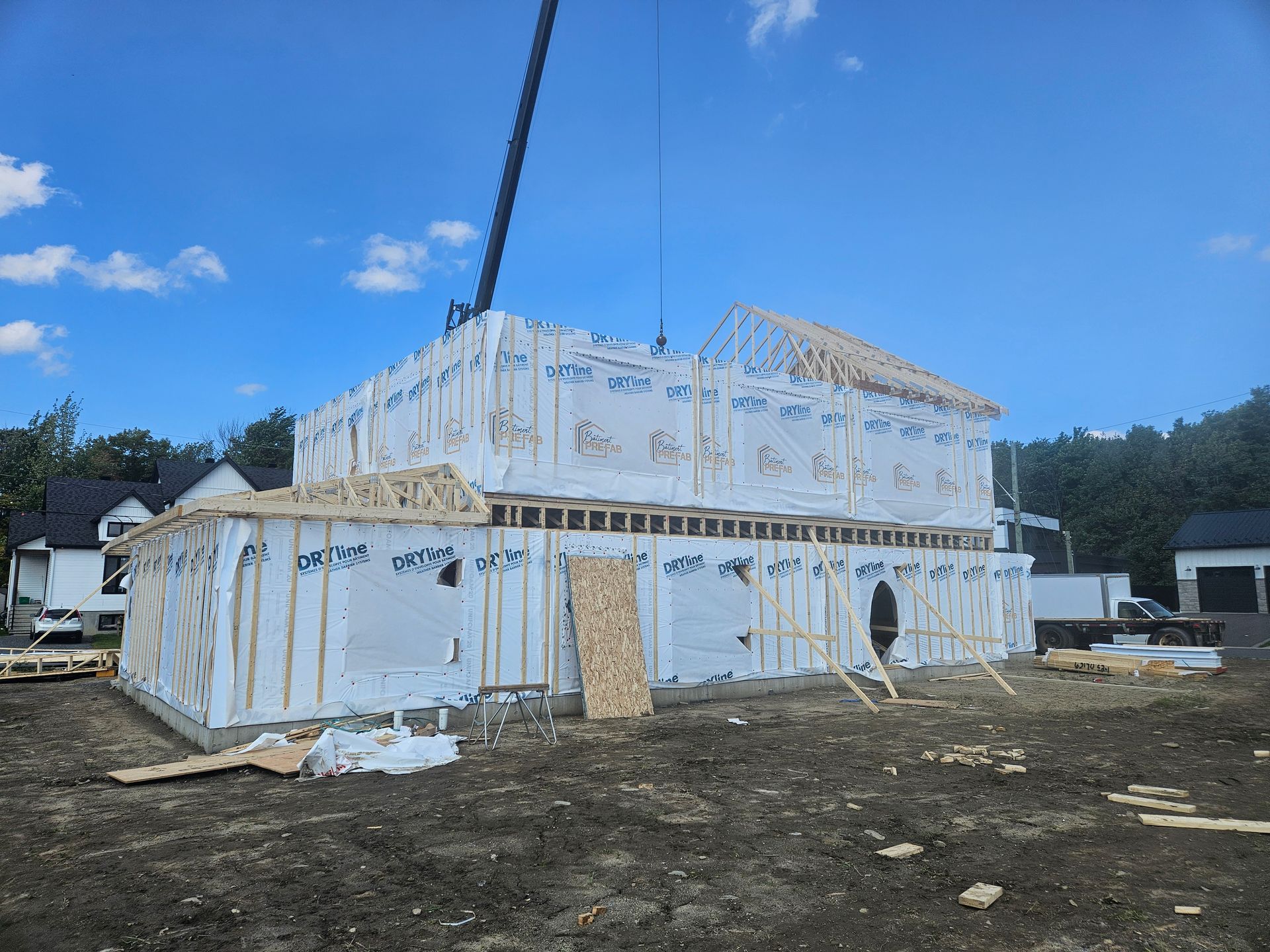 Une maison à ossature bois de deux étages en construction, recouverte d'une bâche blanche, avec une grue visible sur le ciel bleu.