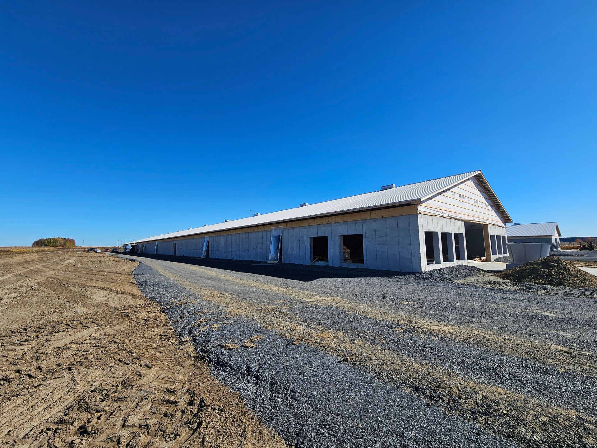 Un long bâtiment agricole de plain-pied, récemment construit, se dresse sous un ciel bleu azur, à côté d'un chemin de gravier et d'un champ de terre.