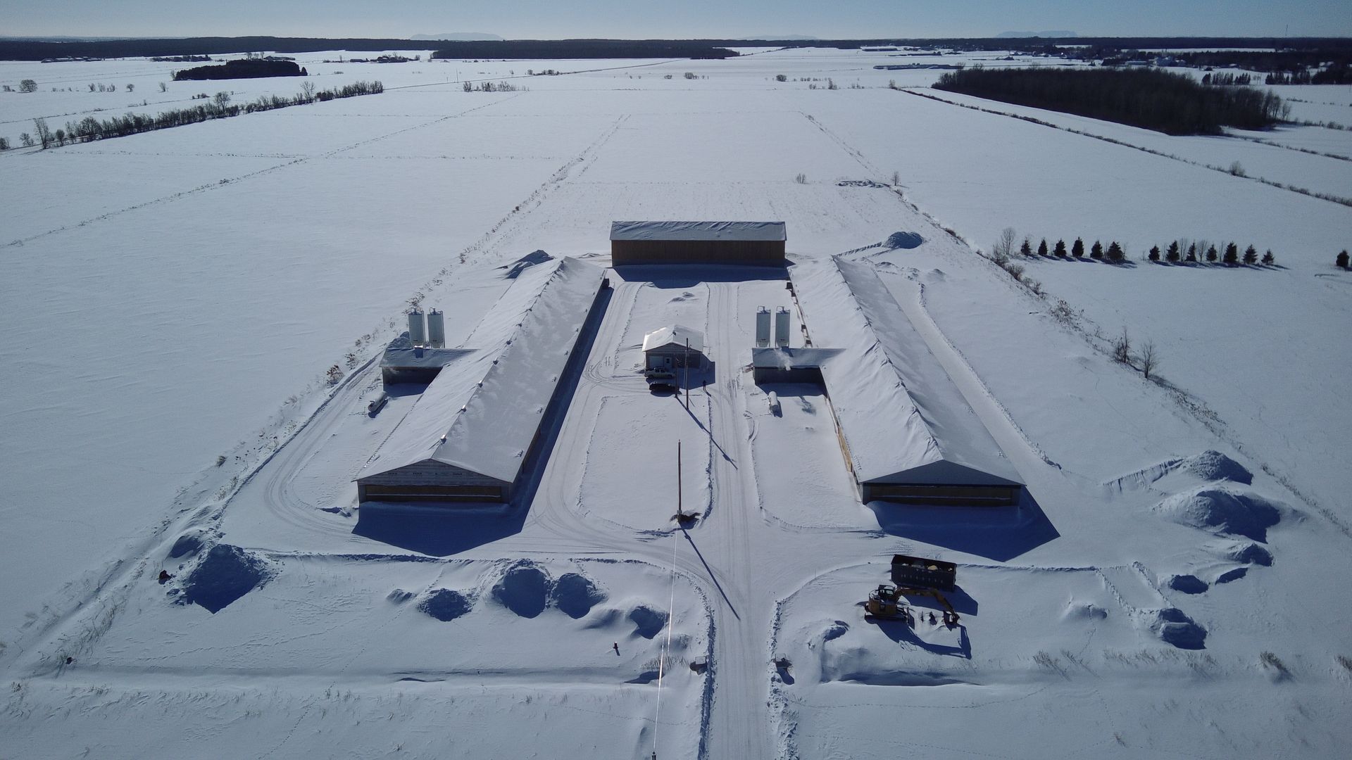 Vue aérienne d'un complexe agricole composé de trois longs bâtiments clairs entourés d'un vaste champ enneigé.