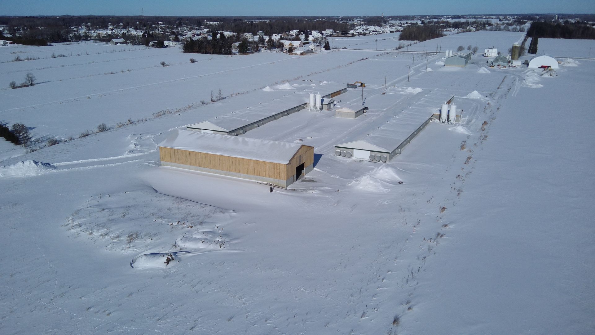 Vue aérienne d'une ferme avec plusieurs longs bâtiments entourés d'une épaisse couche de neige dans un paysage rural.