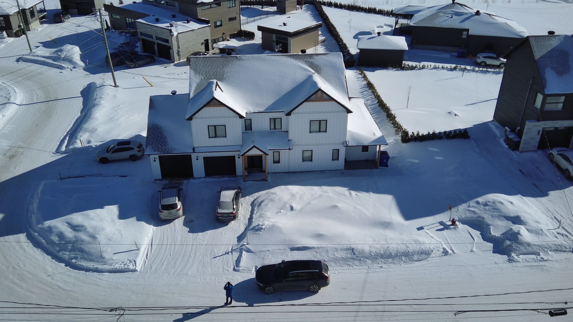 Vue aérienne d'une maison blanche à deux étages entourée d'une épaisse couche de neige, avec plusieurs voitures garées et une allée dégagée.