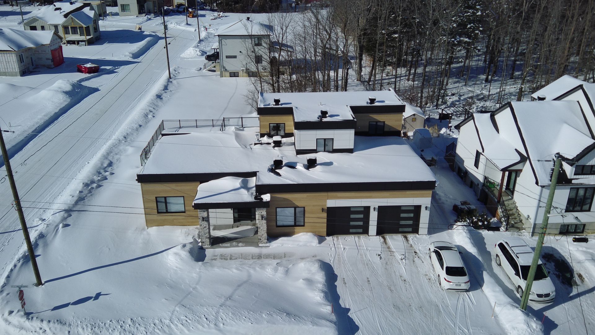 Vue aérienne d'un bâtiment beige à toit plat recouvert de neige, avec un garage double et deux voitures garées devant.