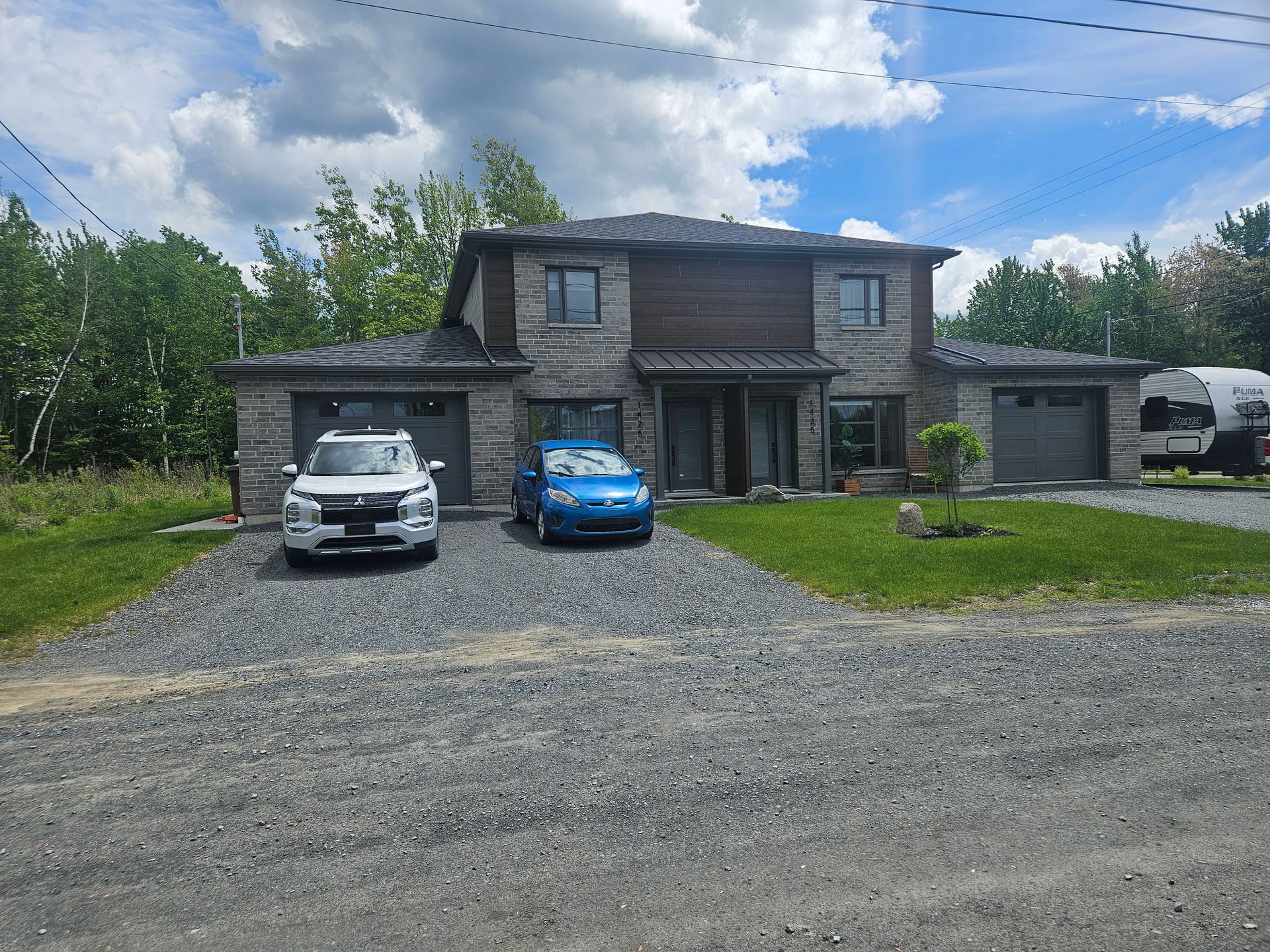 Un duplex moderne de deux étages avec une façade en pierre, deux portes de garage et une allée en gravier avec deux places de stationnement.