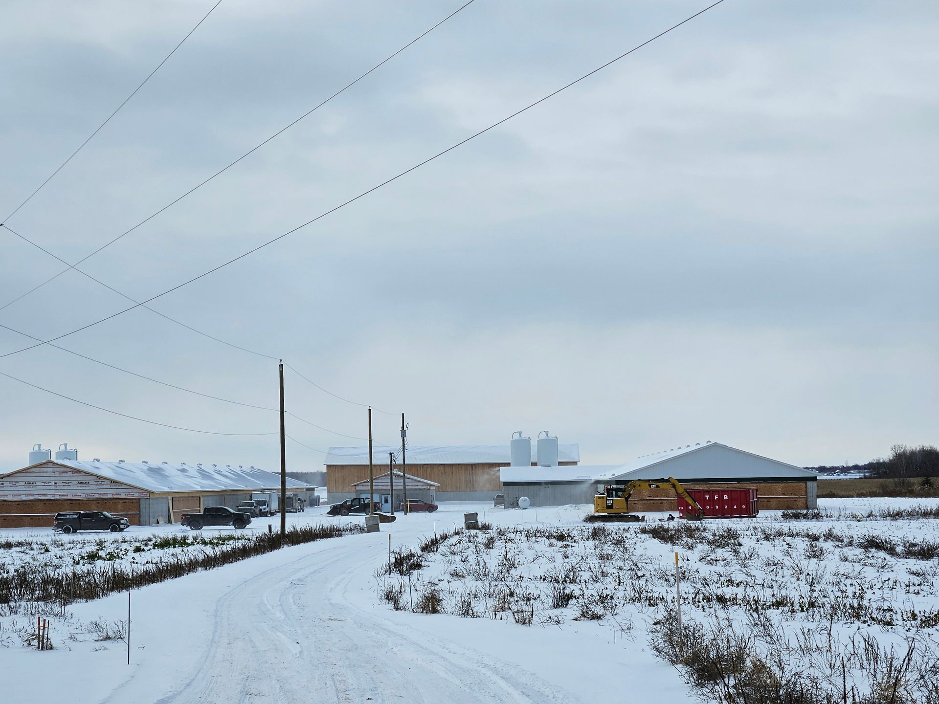 Une scène de ferme enneigée en pleine campagne montre plusieurs longs bâtiments, des silos, des véhicules stationnés et des engins de chantier.