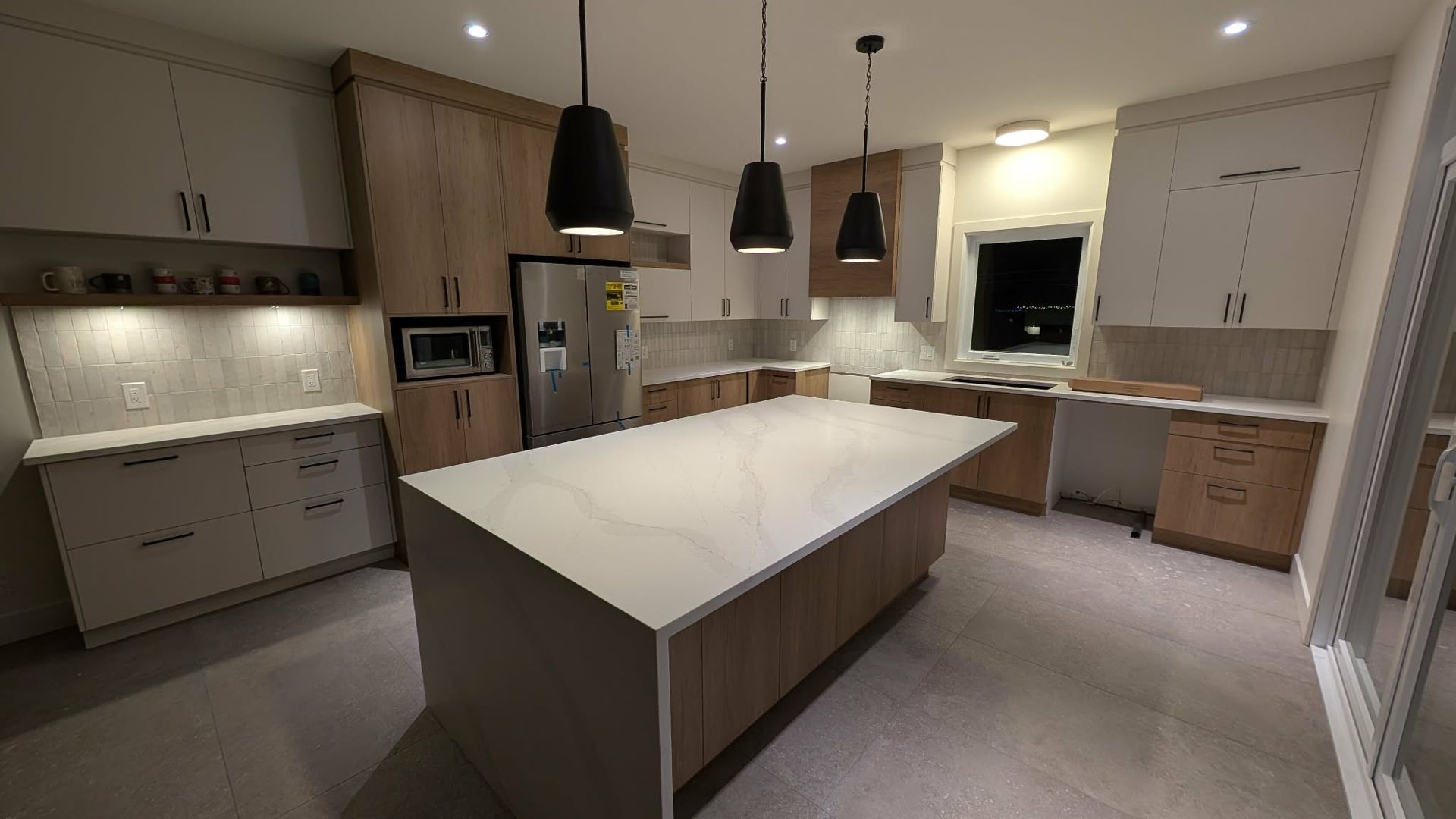 Une cuisine moderne avec un grand îlot central, des armoires en bois clair, des comptoirs blancs et trois suspensions lumineuses.