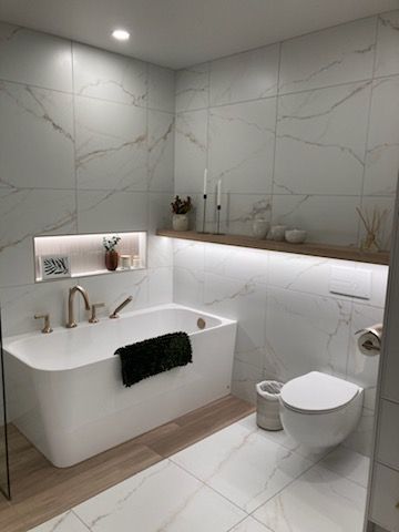 Une salle de bains moderne avec une baignoire blanche, des robinetteries dorées, des toilettes suspendues et des carreaux blancs imitation marbre.