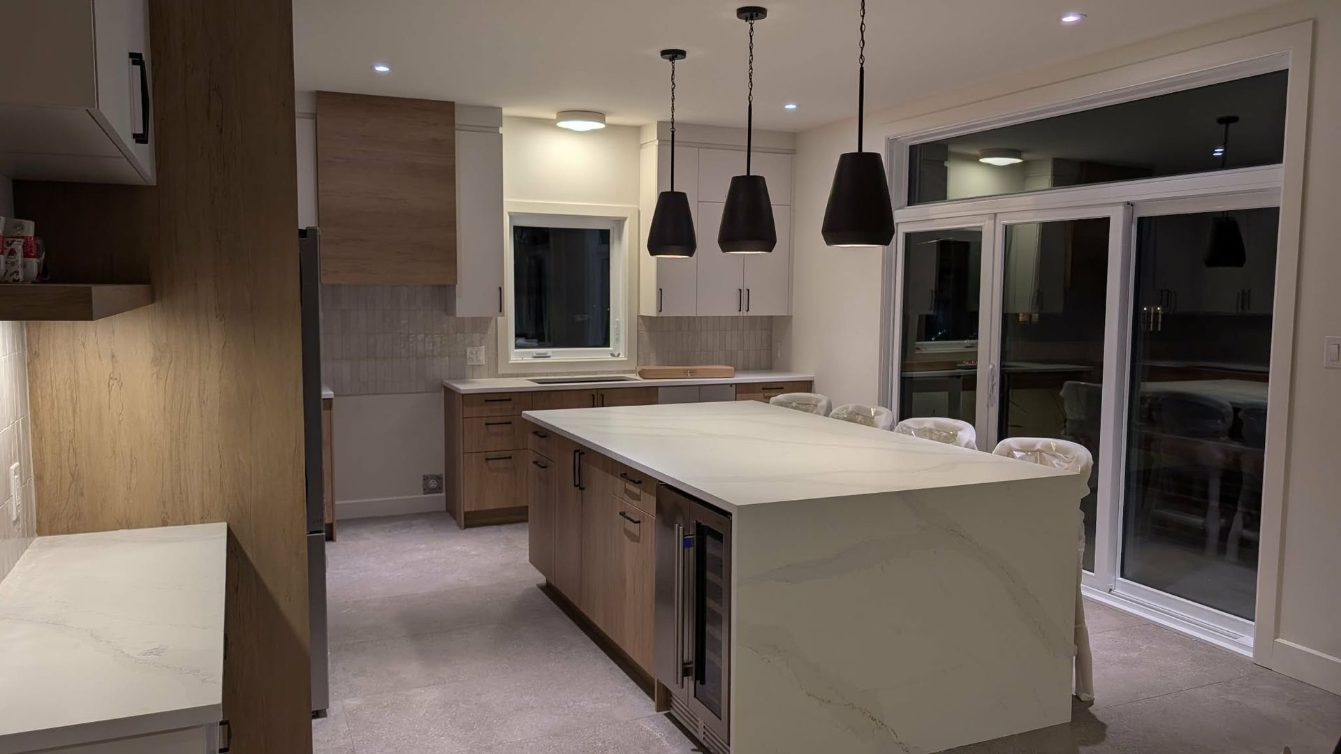 Une cuisine moderne à aire ouverte avec des armoires en bois, un grand îlot en pierre blanche avec des tabourets et des suspensions lumineuses noires.