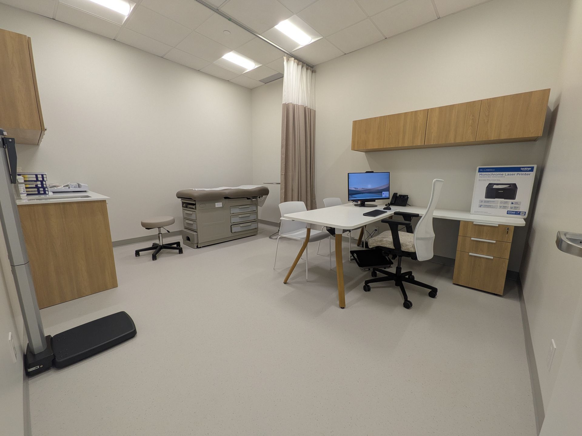 Une salle d'examen médical lumineuse comprenant un bureau avec un ordinateur, une table d'examen, une balance et des meubles en bois clair.