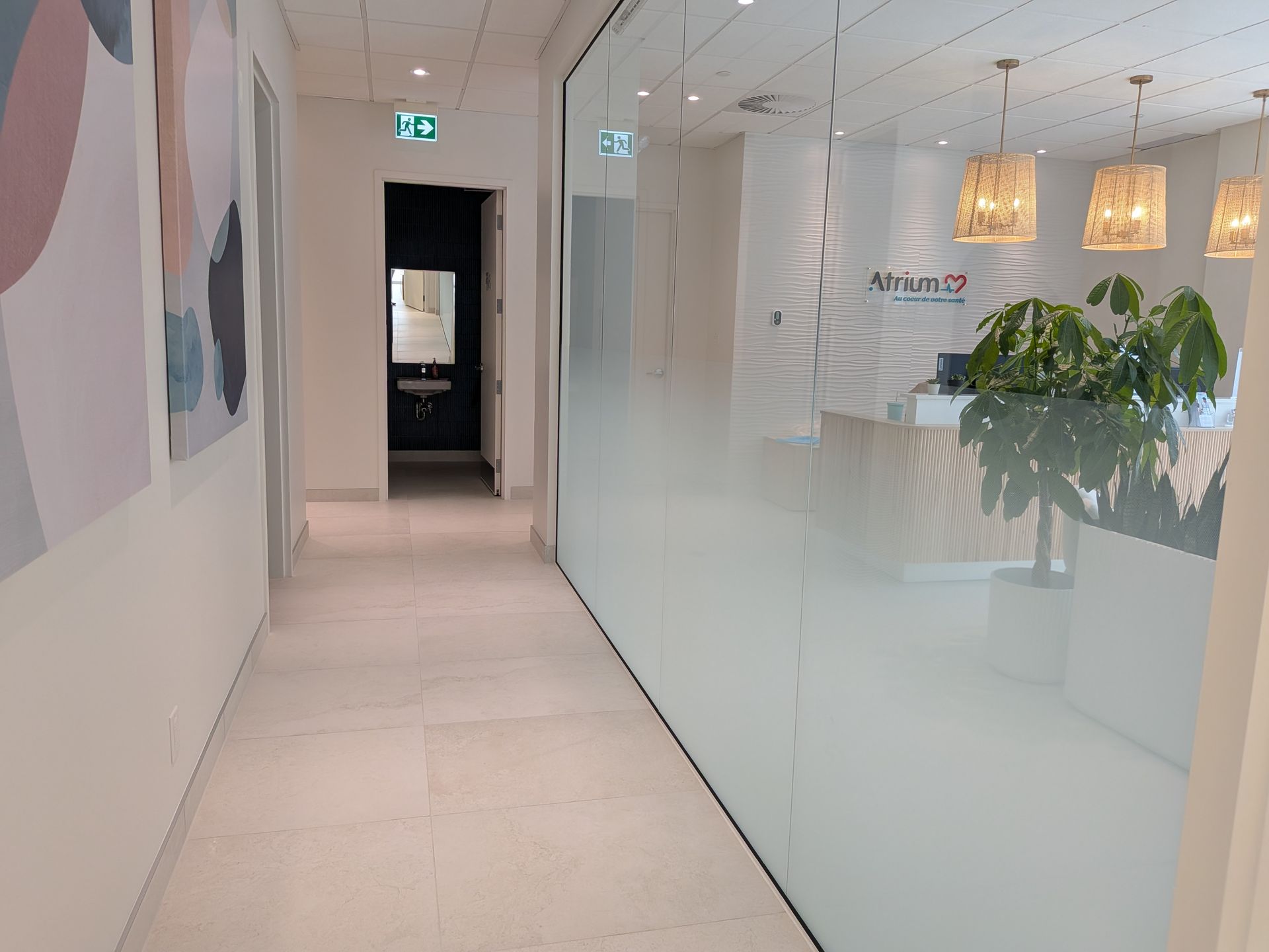 Un couloir de bureau lumineux avec un sol carrelé blanc, une paroi en verre dépoli, des décorations murales et des luminaires suspendus en osier.