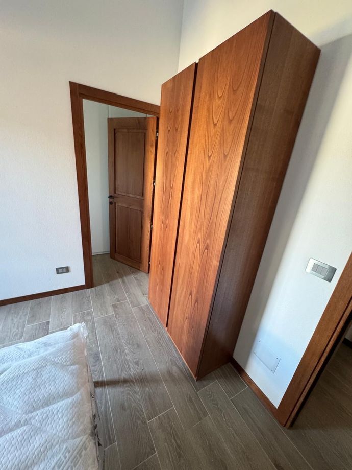 Camera da letto vuota con armadio in legno, porta aperta e pavimento piastrellato vicino al bordo del letto.