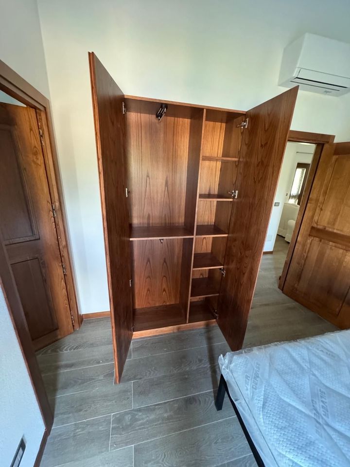 Armadio in legno aperto con ante a specchio in una camera da letto con pavimento grigio e bordo del letto a vista.