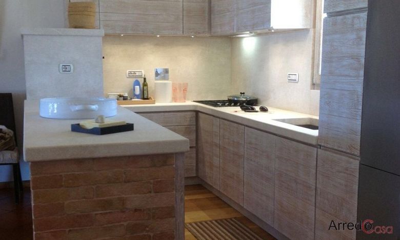 Cucina moderna con mobili in legno chiaro, ripiani bianchi e un'isola piastrellata in primo piano.