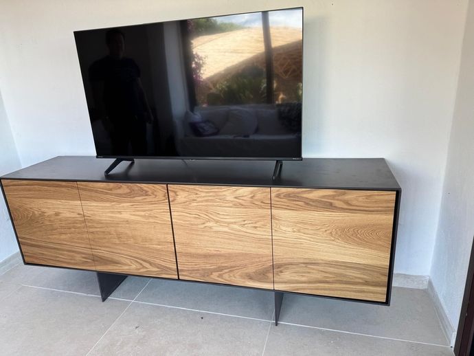 Televisore a schermo piatto su un mobile porta TV con finitura effetto legno in una stanza luminosa con pavimento piastrellato.