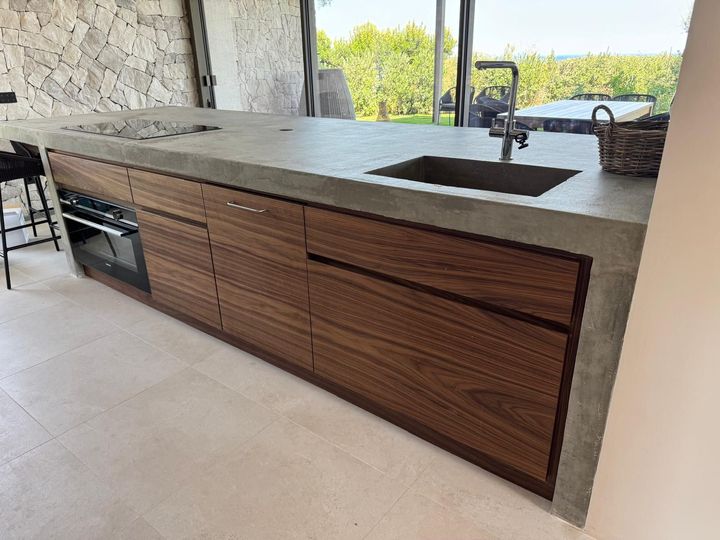 Isola cucina da esterno con mobili in legno, piano di lavoro in pietra, lavello e barbecue integrato.
