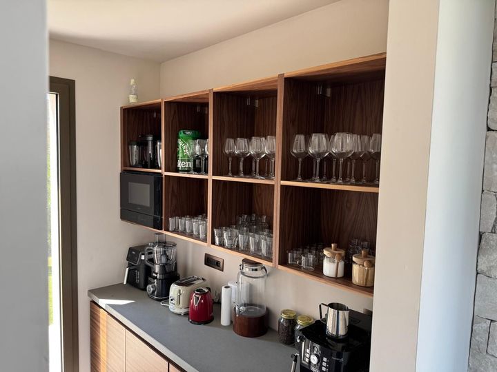 Mobile bar integrato con ripiani in legno, bicchieri, macchine da caffè e piano di lavoro nero in un angolo del corridoio.