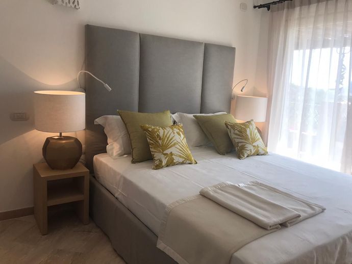 Un letto ben rifatto con testiera grigia, lenzuola bianche, cuscini verdi e lampade da comodino in una camera da letto luminosa.