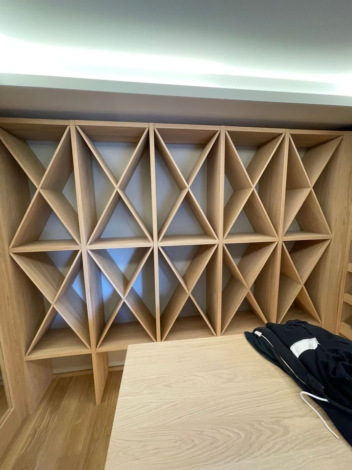 Scaffale geometrico in legno con scomparti triangolari accanto a un tavolo in legno chiaro e una giacca nera