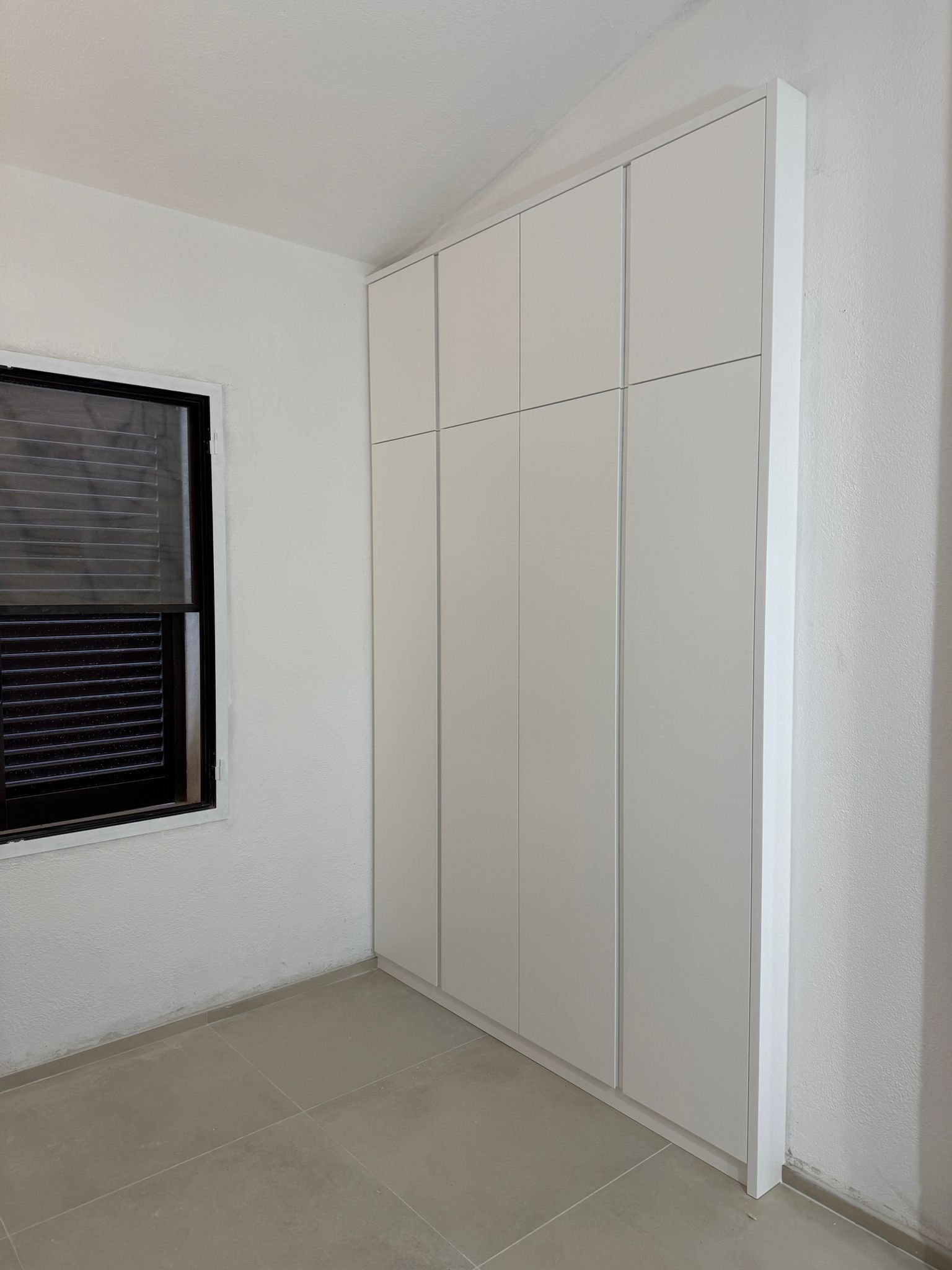 Stanza vuota con ante di armadio a muro bianche, pavimento piastrellato e una finestra con una tenda scura.