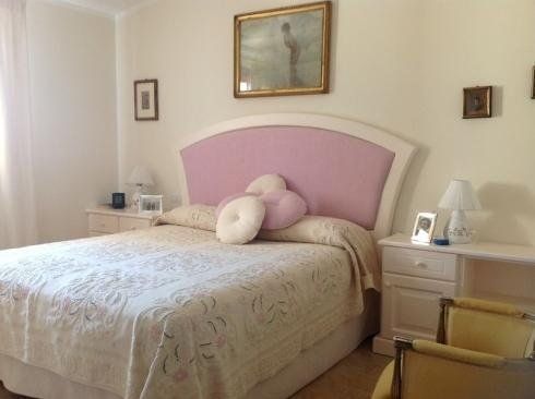 Camera da letto luminosa con letto imbottito rosa, lenzuola bianche, comodini e quadri alle pareti.