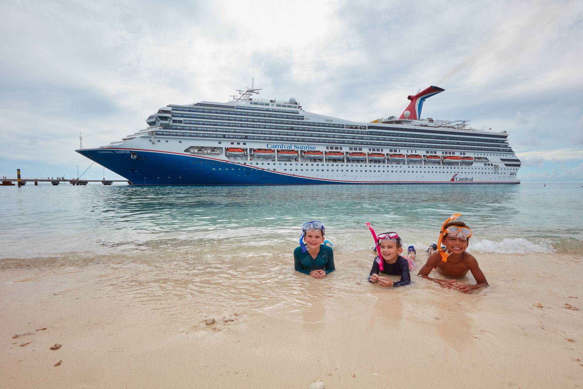 Three people with snorkeling masks lie in shallow tropical water with a large cruise ship docked in the background.