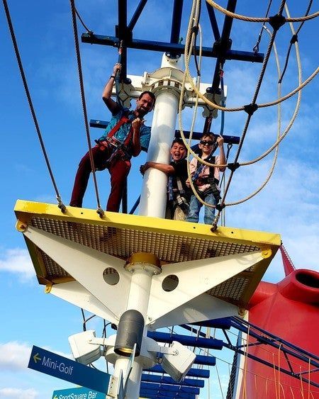 People climbing a pirate-ship ropes course against a blue sky