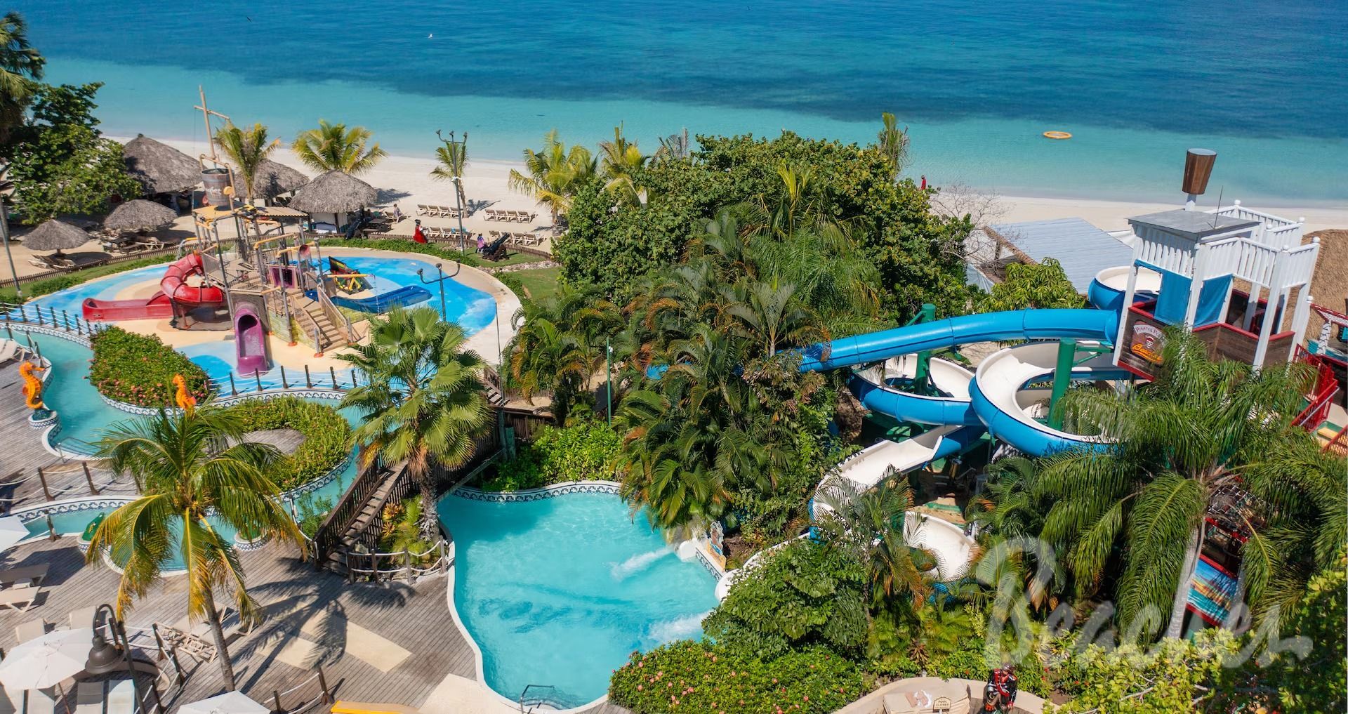 Aerial view of a tropical beach resort with water slides, a swimming pool, and a children's play area near the ocean.