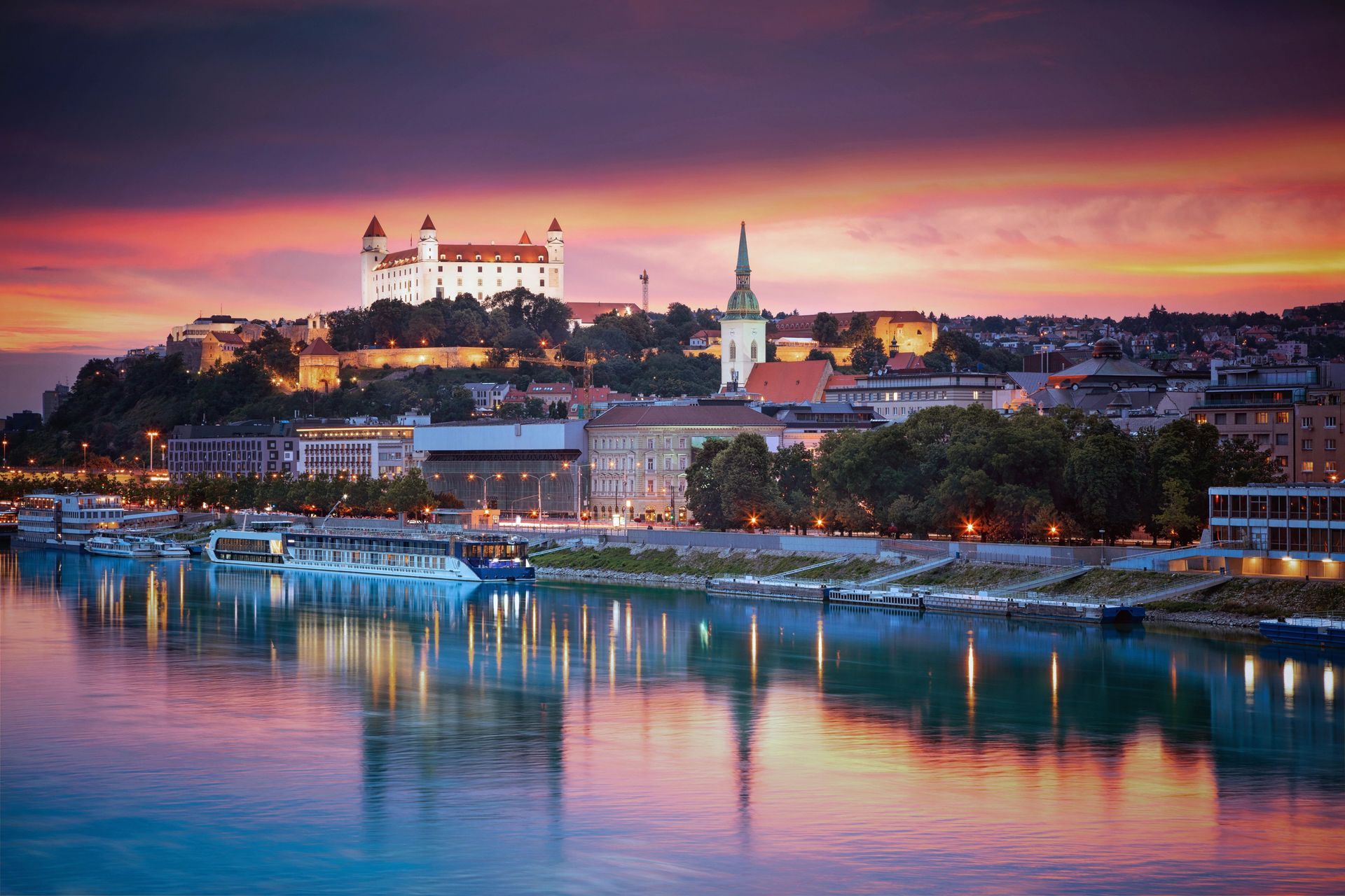 Sunset over a riverside city with a lit castle on the hill and colorful reflections on the water