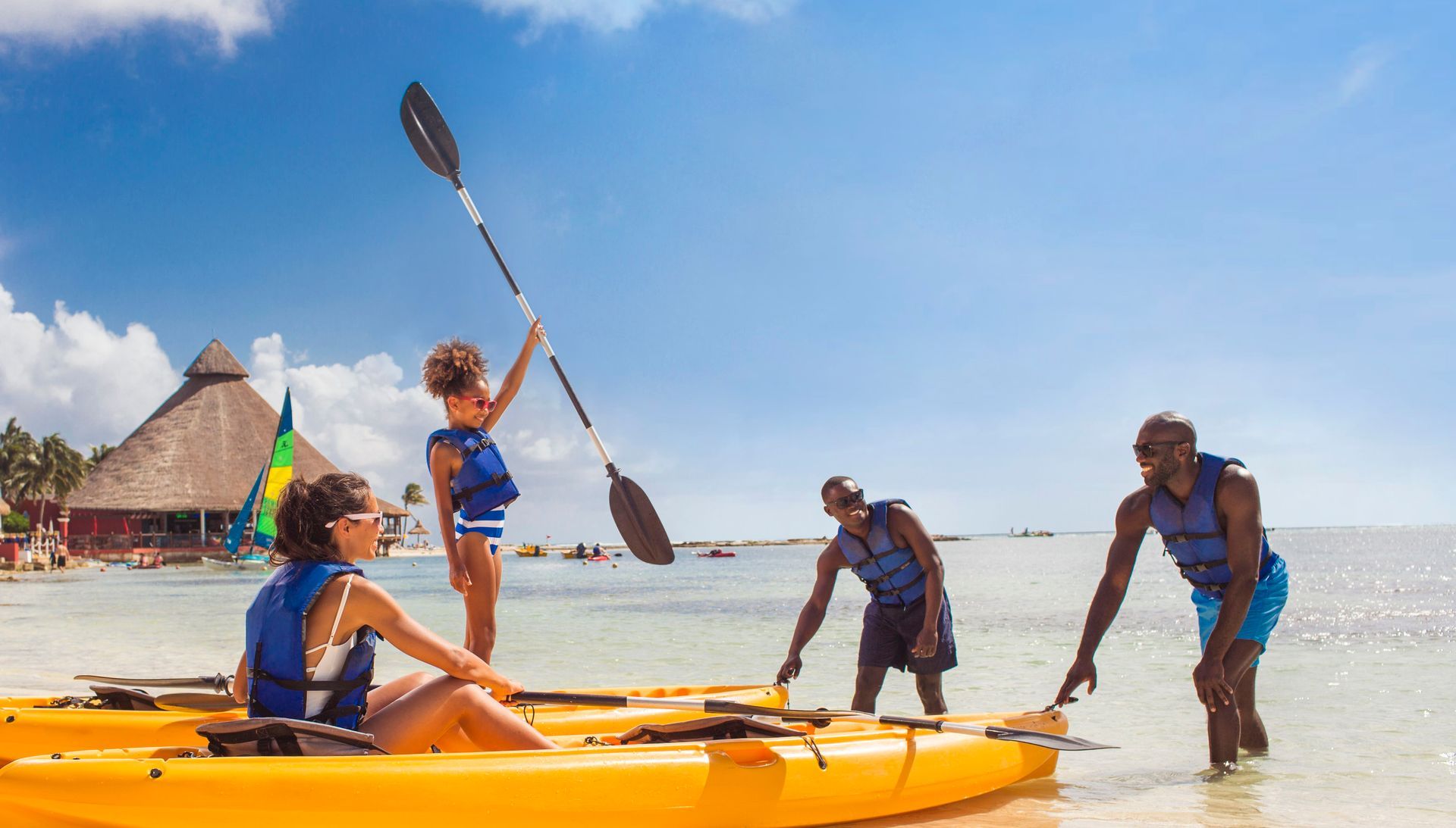 People on kayaks and a small sailboat playing in shallow tropical water near a sandy beach.