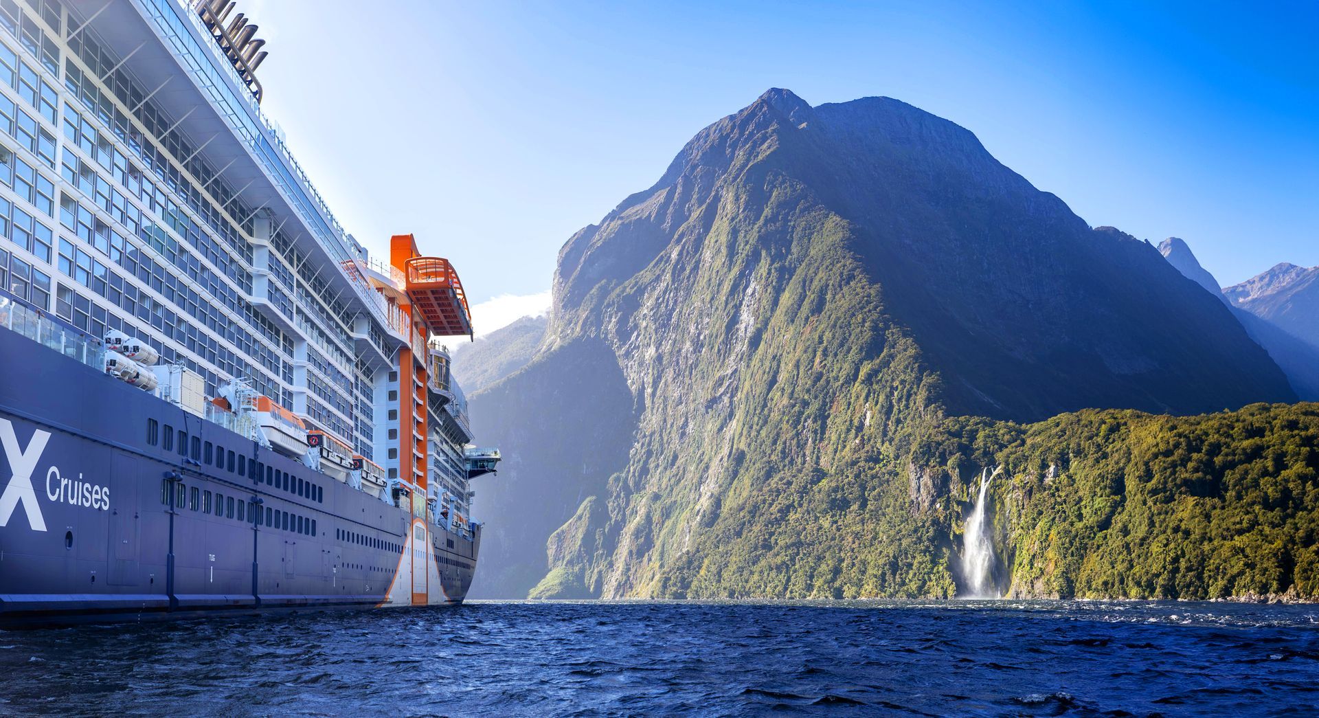 Cruise ship sailing beside a steep green mountain and blue water under a clear sky