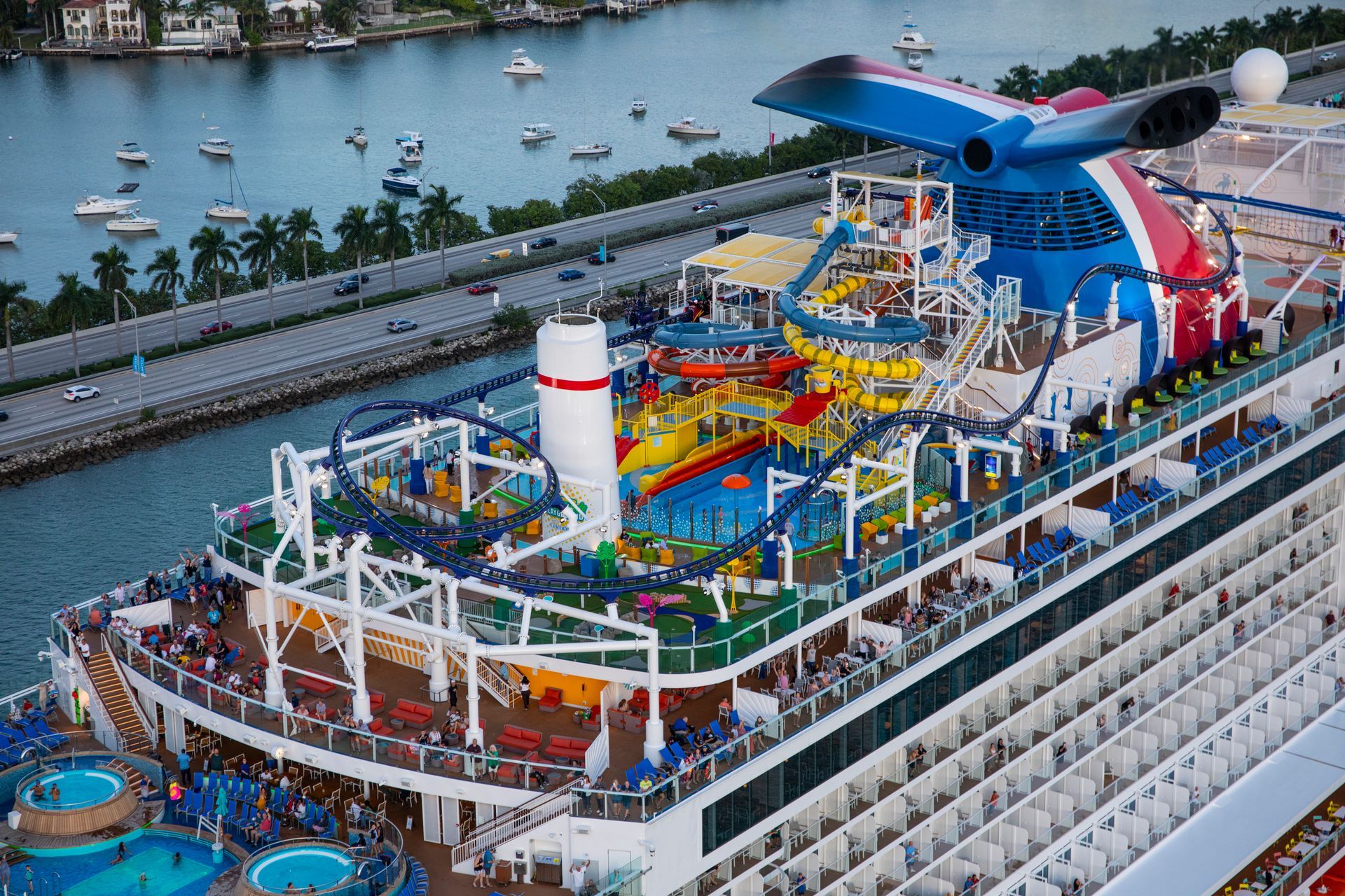 Cruise ship deck with colorful water slides, pool, and lounging area beside a calm harbor