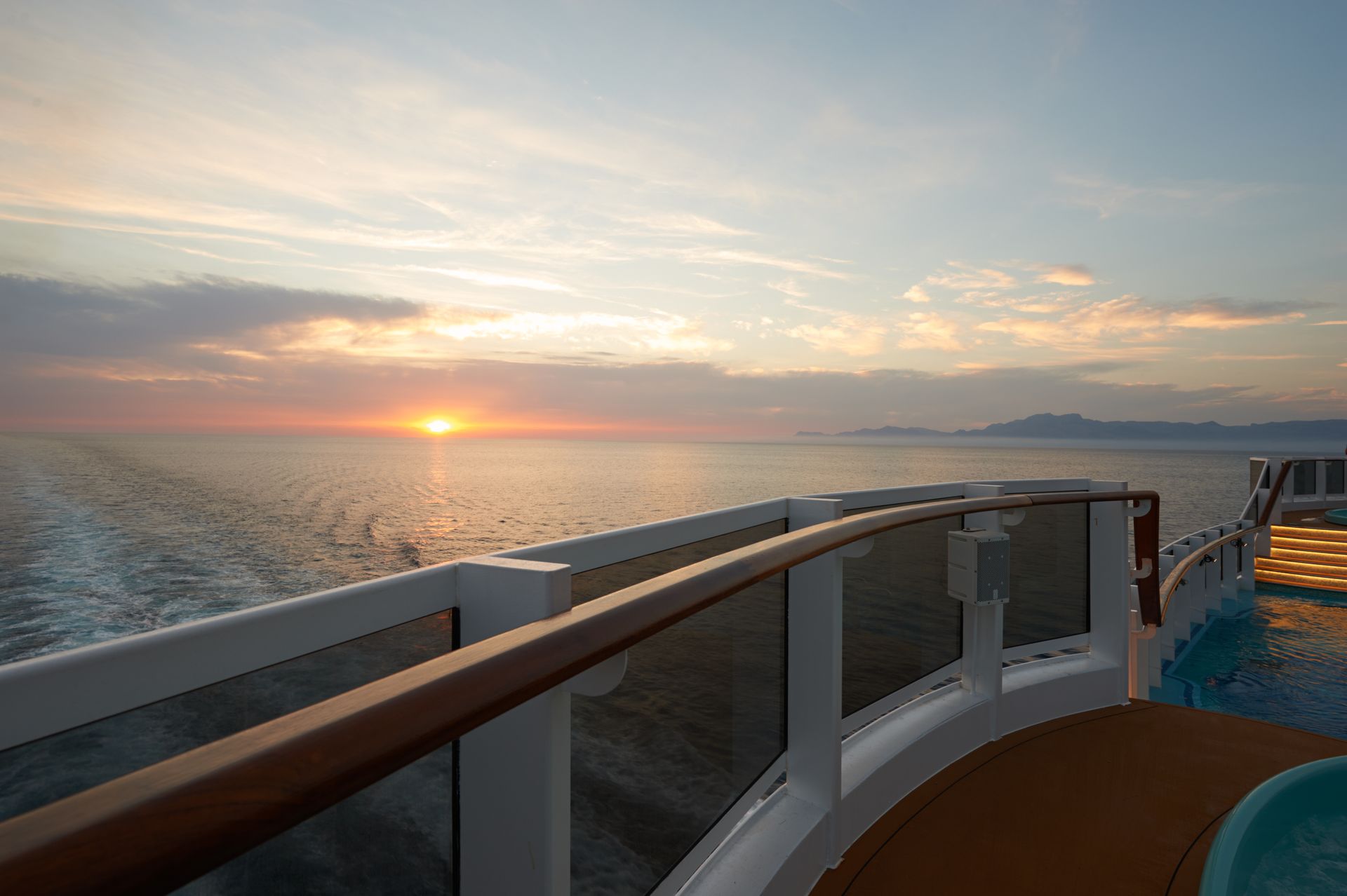 Sunset over the ocean from a ship’s deck, with white railings and calm water.