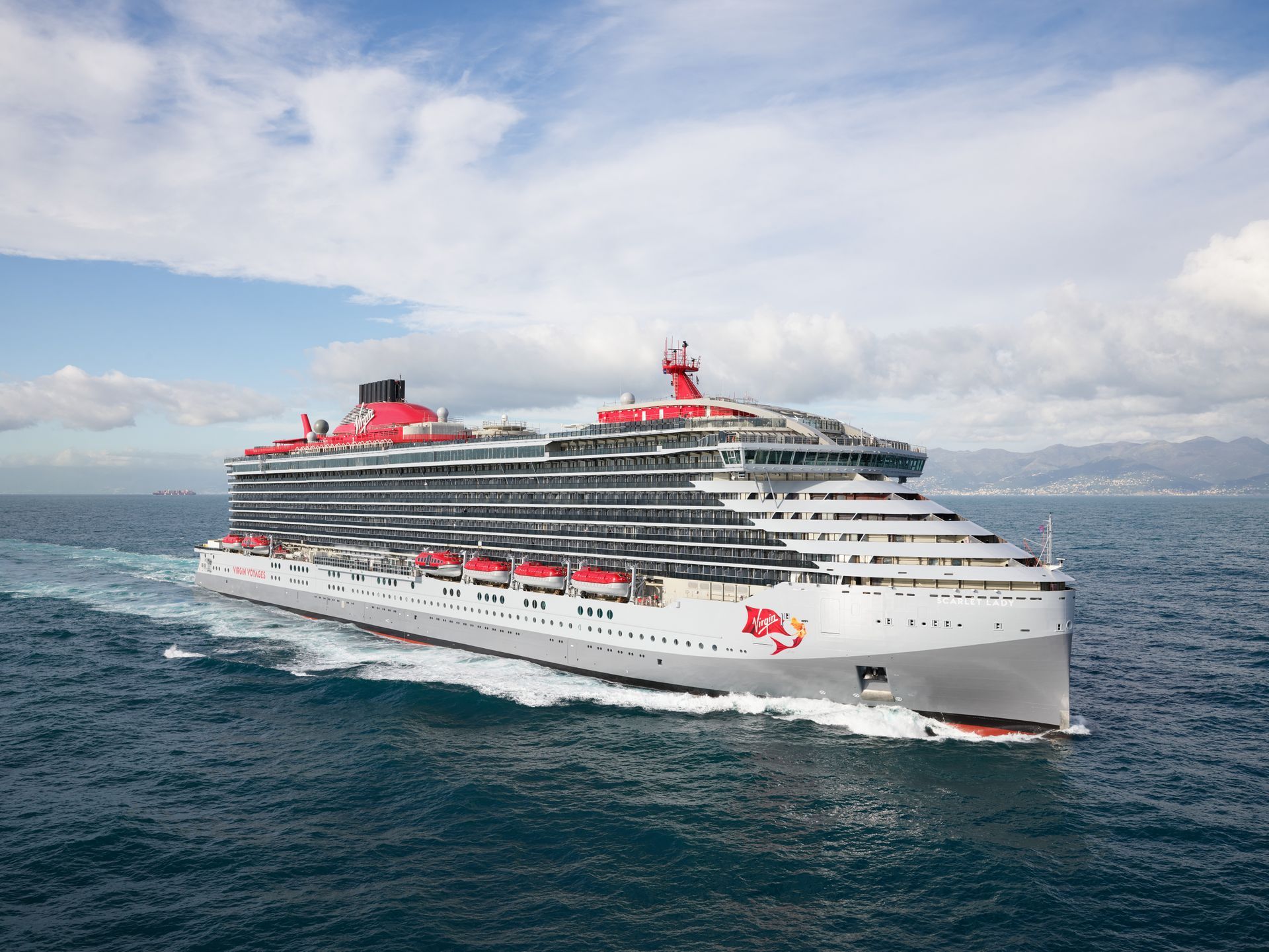 A white Virgin Voyages cruise ship with red accents sailing on blue water under a cloudy sky.