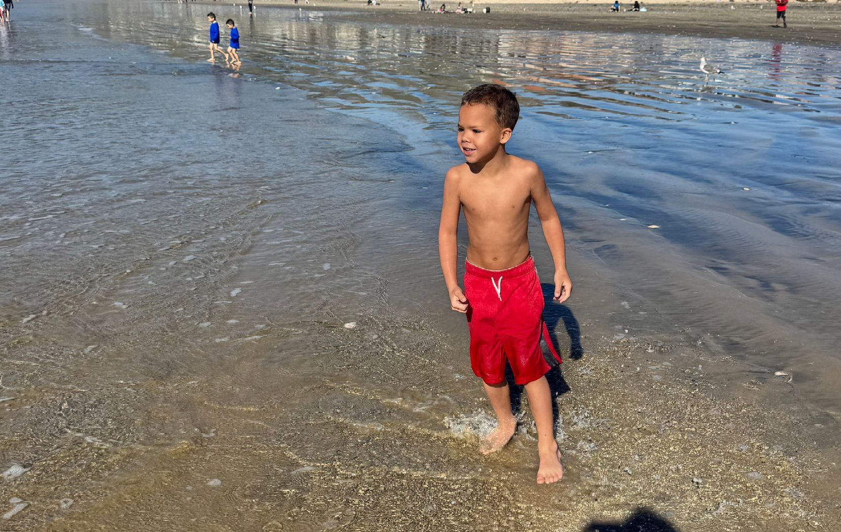 A child stands in shallow beach water wearing red swim trunks, looking toward the side under a clear blue sky.