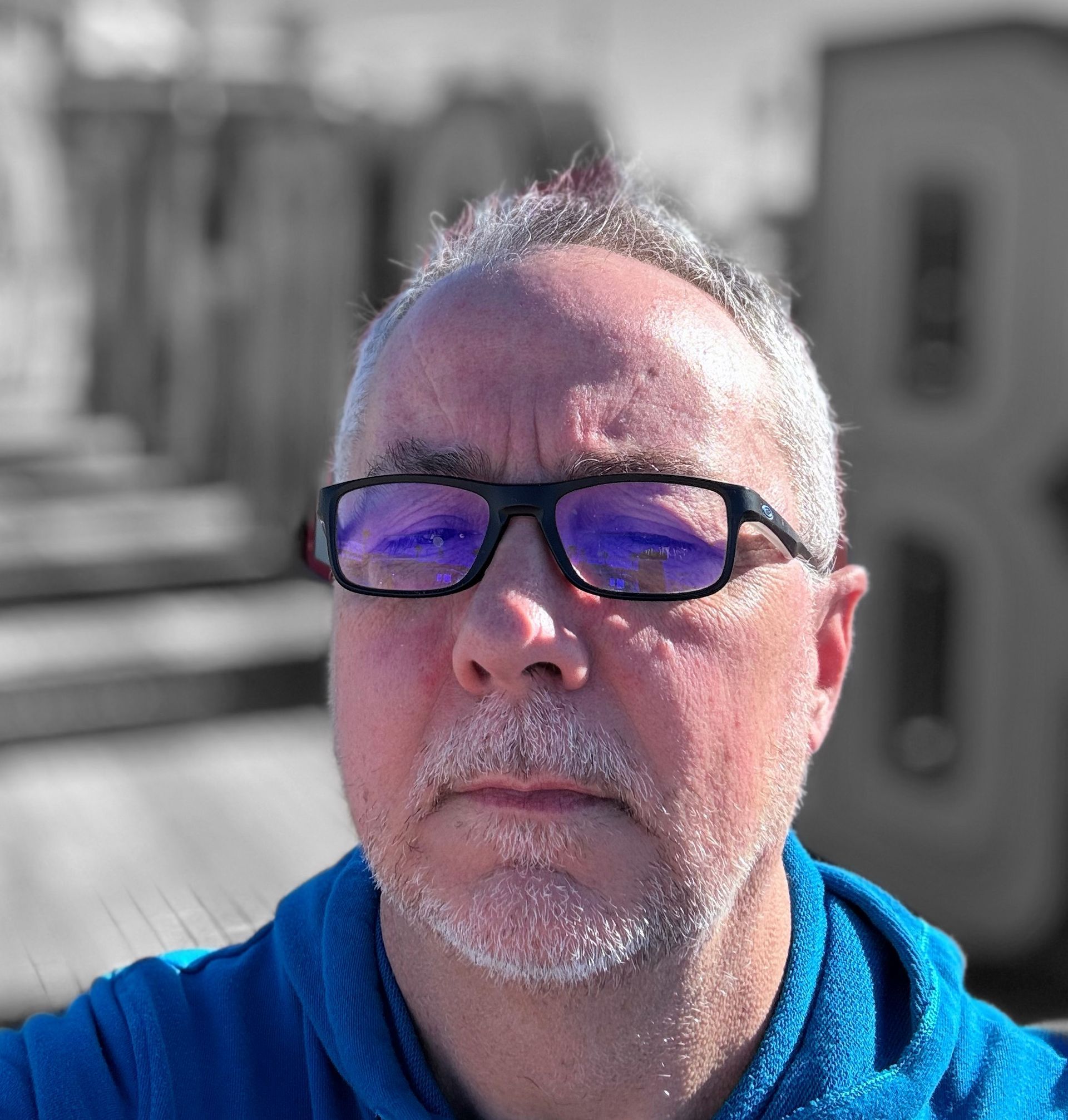 A selfie of a person with short hair, wearing blue-tinted glasses and a blue hoodie, against a blurred background.