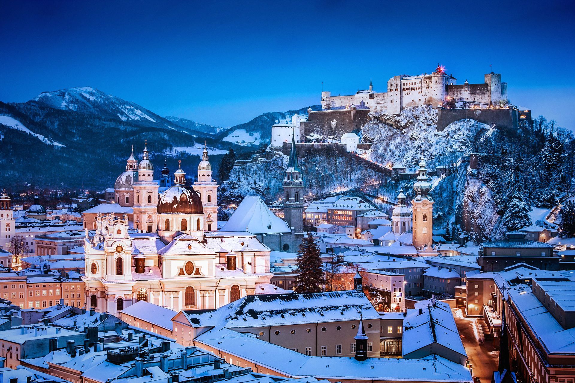 Snowy European town at dusk with a lit church in the foreground and a hilltop castle above.