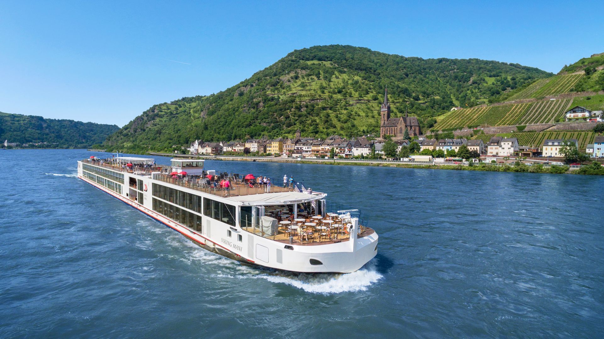 A white river cruise ship travels on a blue river past a hillside town with vineyards and a church.