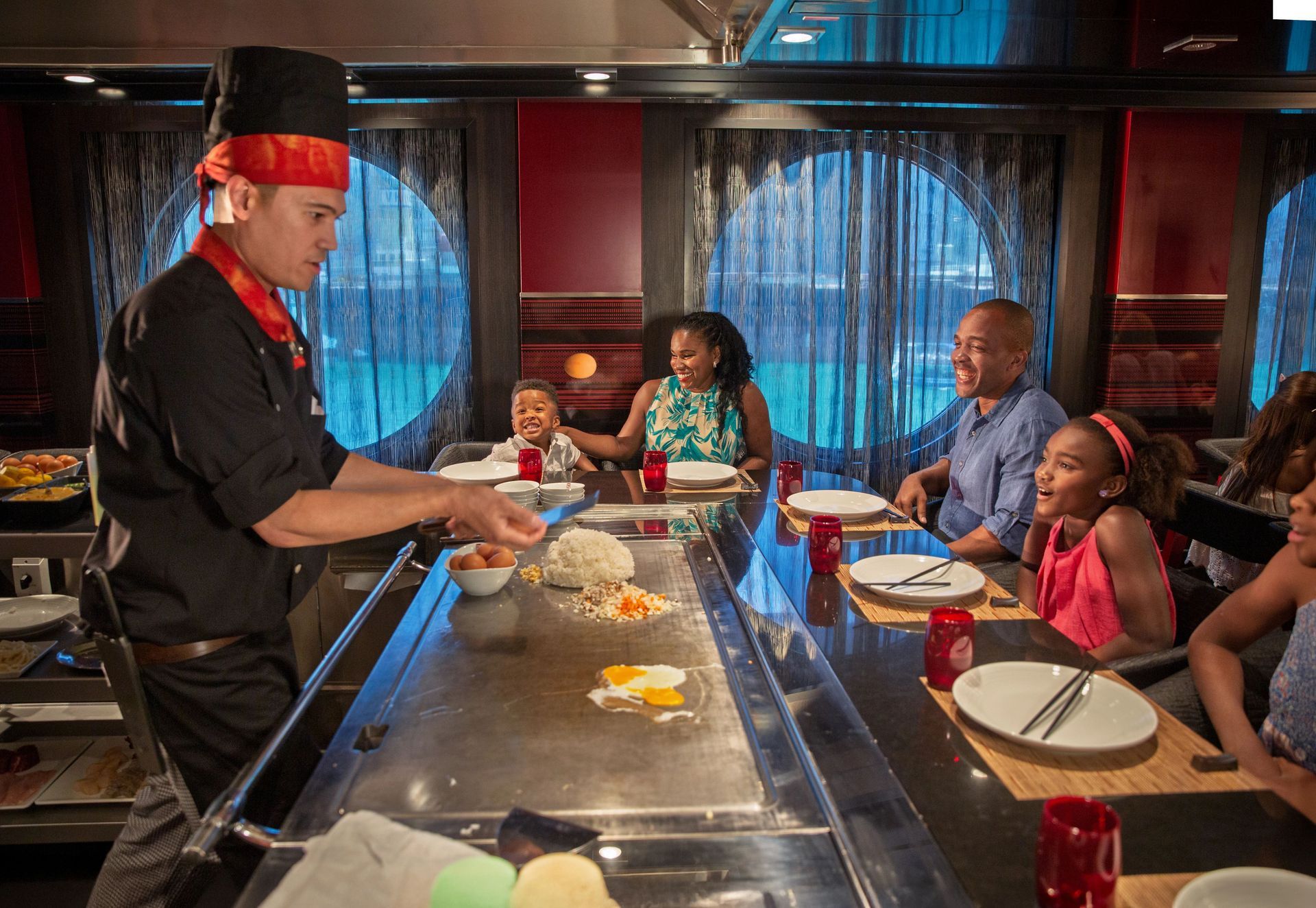 A chef in a black uniform prepares food on a teppanyaki grill for a family seated at a dining table.