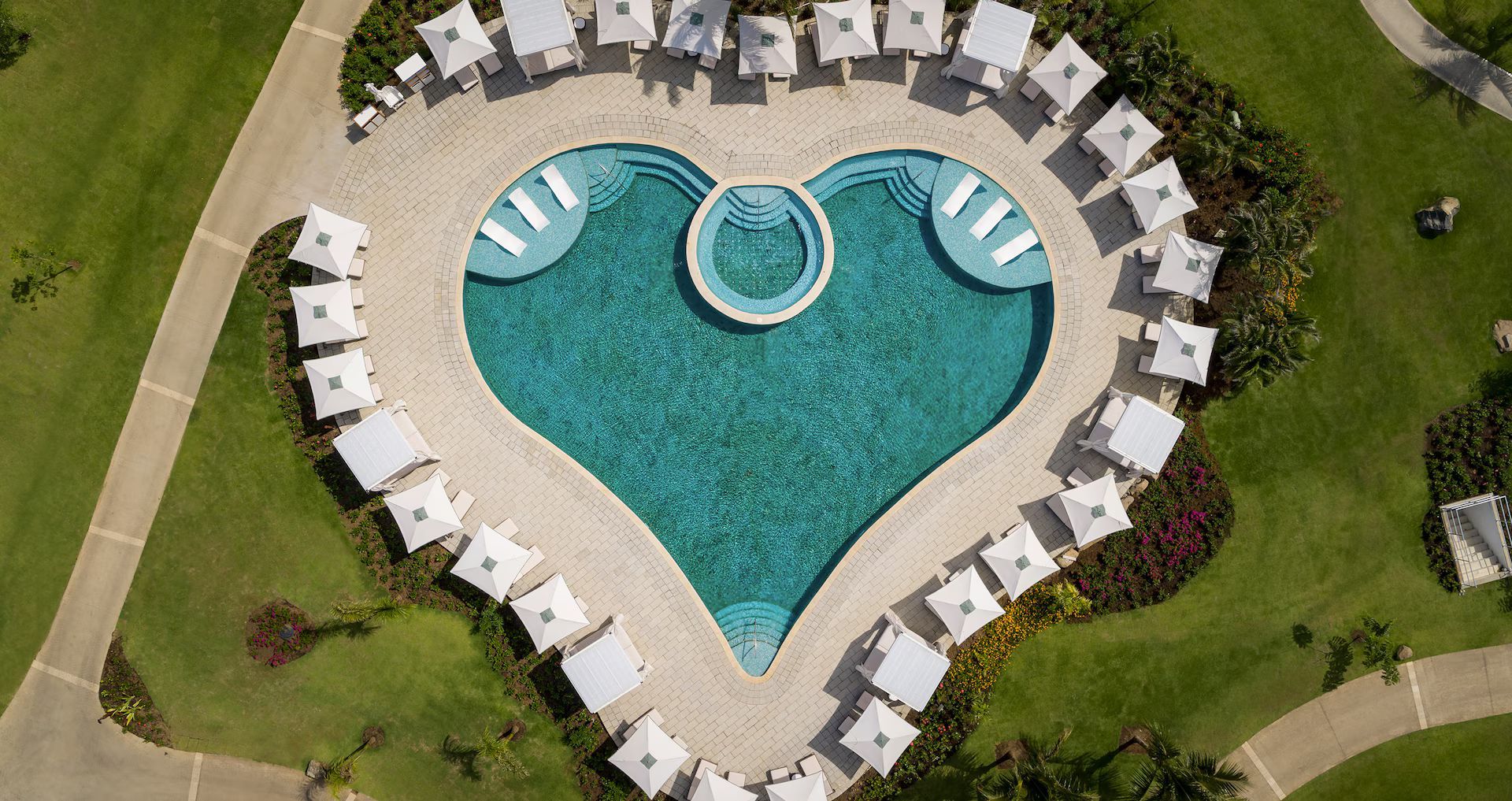 An aerial view of a heart-shaped turquoise swimming pool surrounded by white lounge chairs on a paved patio and grass.