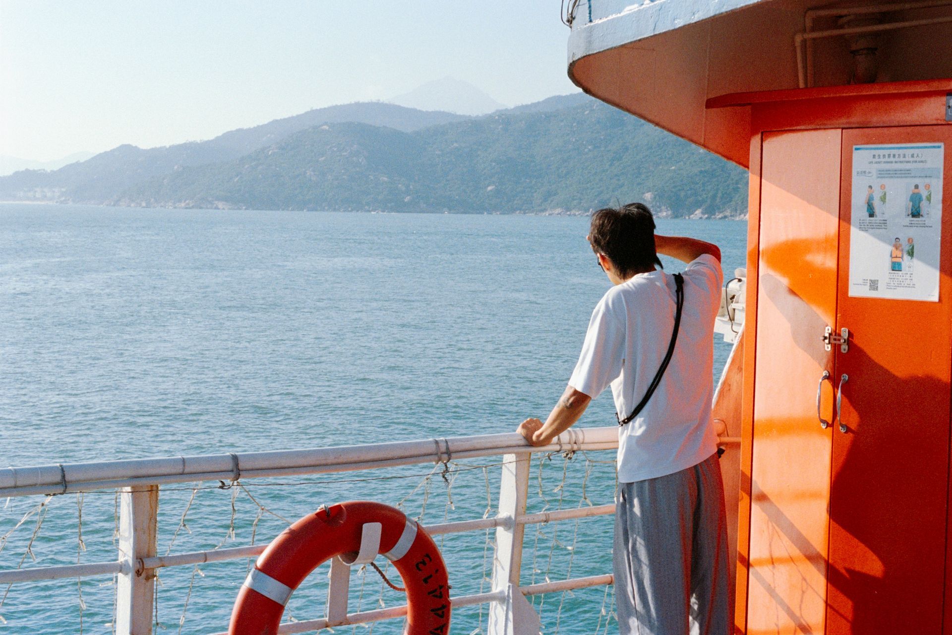 Person on a ferry deck looks over the sea toward distant mountains beside an orange wall.