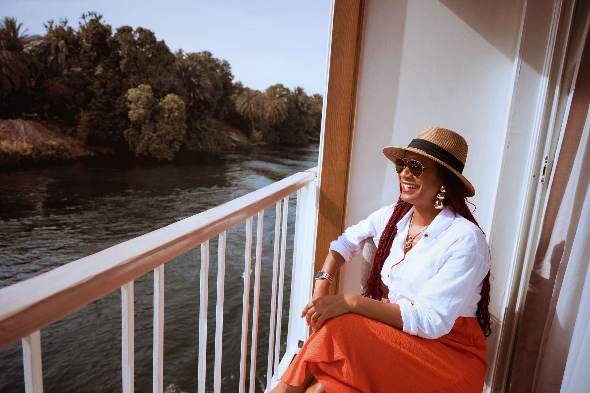 Woman in a straw hat and orange skirt sitting on a balcony by a river, smiling in the sun.