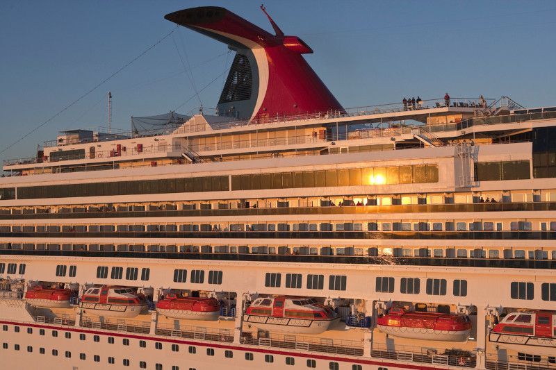 A large white Carnival cruise ship with its signature red, wing-shaped funnel against a clear sky at sunset.