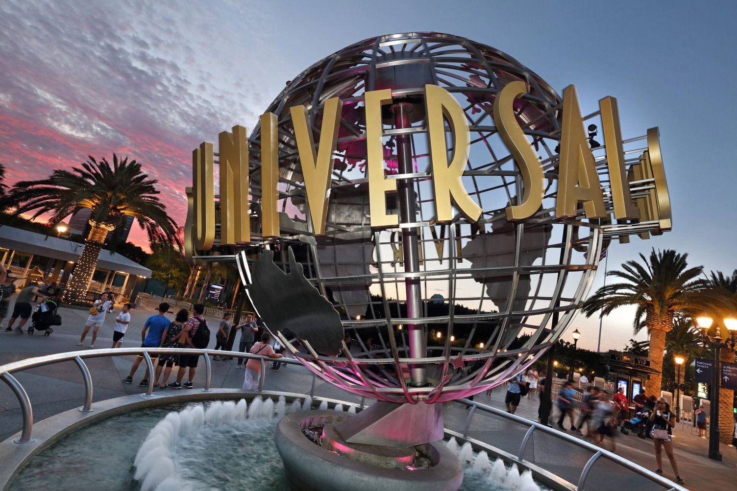 The iconic Universal Studios rotating globe sign at sunset, with a fountain in the foreground and park visitors nearby.